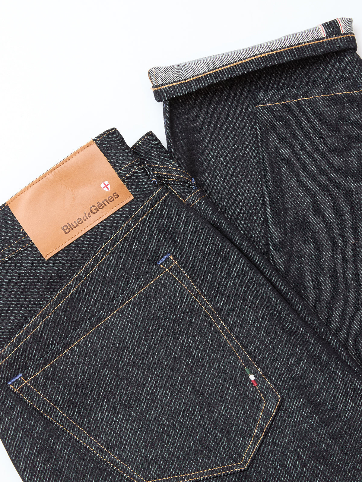 The Blue de Gênes Giacomo Selvedge V2 Jeans in raw denim feature dark blue rigid fabric, tan stitching, a branded leather patch, and a rolled-up leg to reveal the selvedge edge. Designed in a relaxed loose fit for comfort.