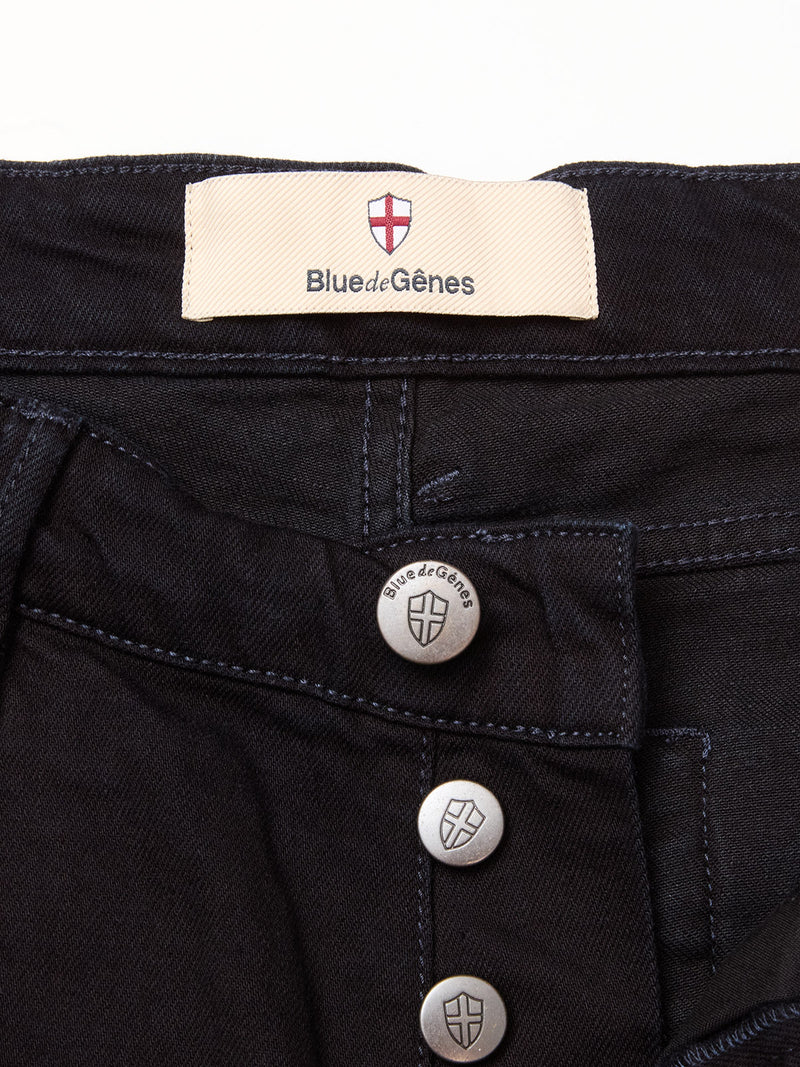 Close-up of Vinci Venom Blk Jeans - Dark Denim by Blue de Gênes, featuring black comfort stretch twill, three exposed silver shield-engraved buttons with the brand name, and a cream label with the brand logo stitched inside the waistband.