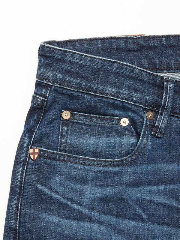 Close-up of Vinci Used Jeans - Used by Blue de Gênes in blue comfort stretch denim, showing the waistband, front pocket with metallic button, coin pocket, and shield-shaped logo near the pocket. Made in Italy.