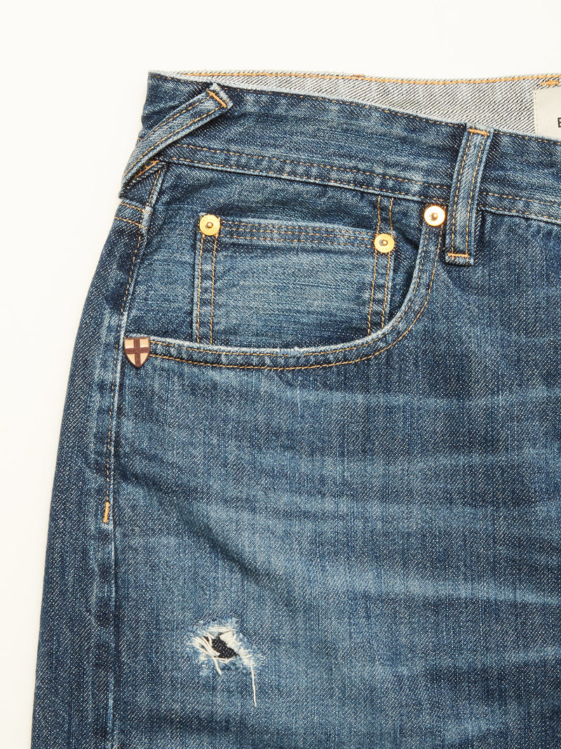 Close-up of Blue de Gênes Giacomo Osaka Jeans in dark denim, featuring yellow stitching, a small front pocket, a distressed thigh rip, and a tiny shield-shaped emblem near the pocket.
