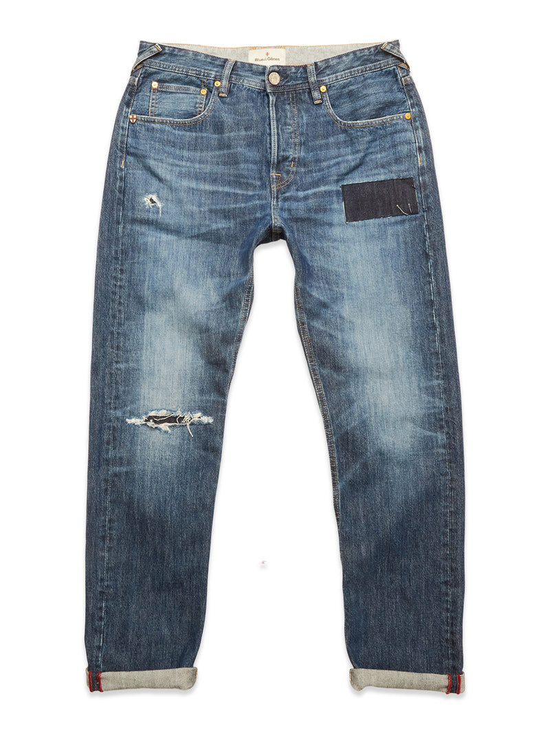 The Giacomo Osaka Jeans - Dark Denim by Blue de Gênes feature rigid blue denim, a faded wash, rolled cuffs, distressed details, a left thigh tear, and a dark patch on the right thigh. Made in Italy.