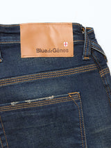 Close-up of the back pocket and waistband of Blue de Gênes Repi Dark Vintage Jeans in mid blue denim, with a brown leather brand patch and small red-white crest on comfort stretch denim.