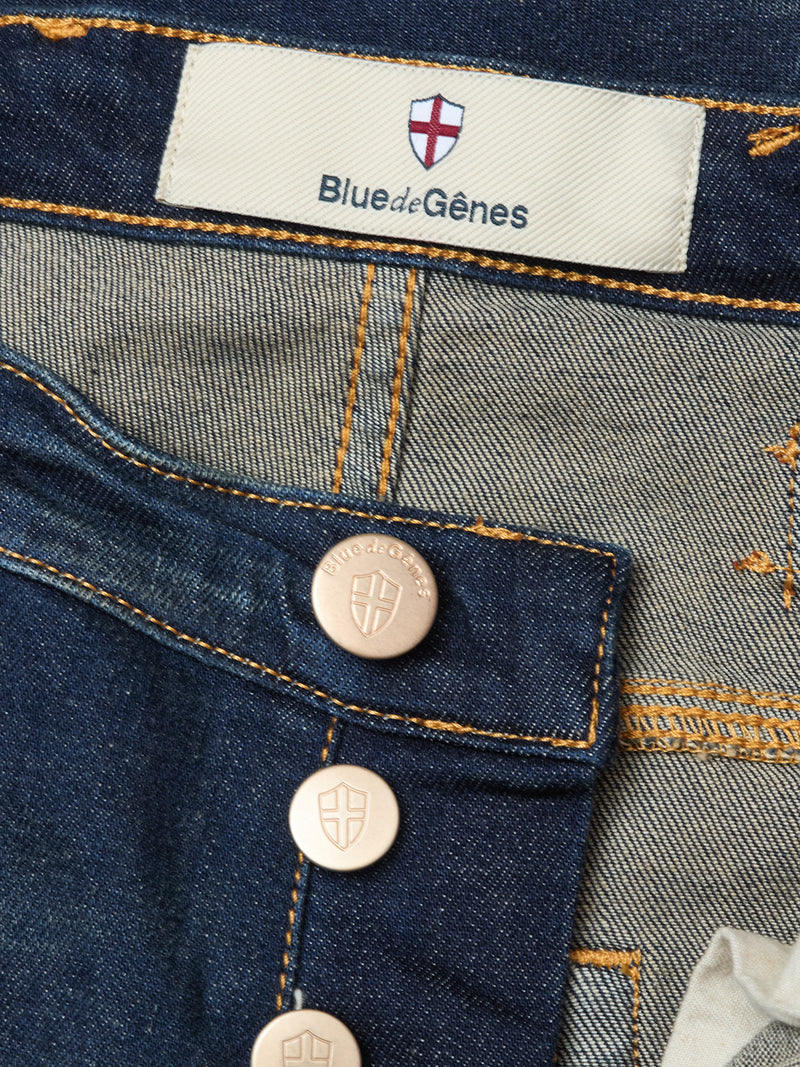 Close-up of Blue de Gênes Repi Dark Vintage Jeans in mid-blue denim, featuring a regular slim fit, visible buttons and stitching, comfort stretch fabric, and a red cross emblem on the label above the waistband.