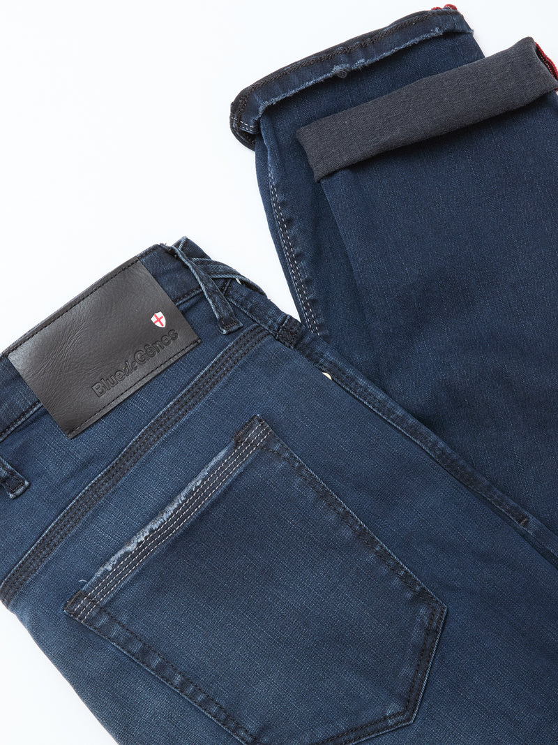 The Repi BlueBlk Used Jeans by Blue de Gênes feature a dark blue-black slim fit, laid flat with one leg folded. Details include a back pocket, belt loops, and a black leather patch with a red and white shield logo. Made from comfort stretch denim.