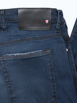 Close-up of the Repi BlueBlk Used Jeans by Blue de Gênes in blue-black denim, featuring a leather waistband patch with the brand logo and a red-and-white shield emblem above the right back pocket. Made from comfort stretch denim for style and ease.