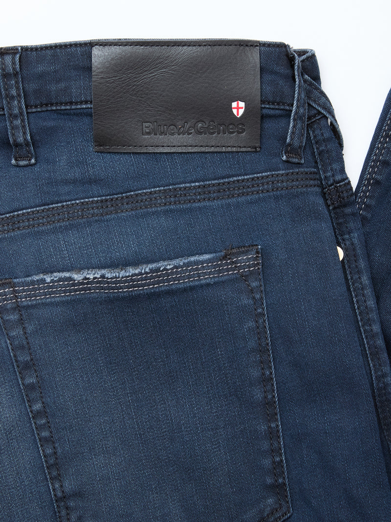 Close-up of the Repi BlueBlk Used Jeans by Blue de Gênes in blue-black denim, featuring a leather waistband patch with the brand logo and a red-and-white shield emblem above the right back pocket. Made from comfort stretch denim for style and ease.