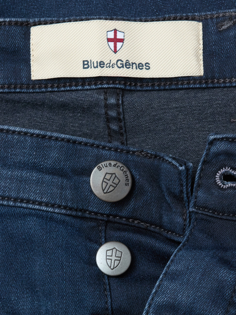 Close-up of Blue de Gênes Repi BlueBlk Used Jeans in blue-black stretch denim, with two shield-embossed metal buttons and a label above the waistband featuring a red cross and the Blue de Gênes brand name.