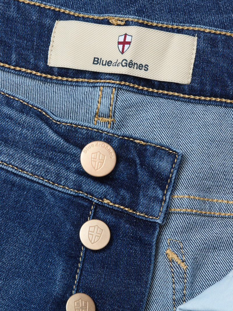 Close-up of Blue de Gênes Recco Blue Vintage Jeans in vintage wash, highlighting comfort denim, visible brass buttons, red and white shield logo label, detailed stitching, rich denim texture, and tapered legs.