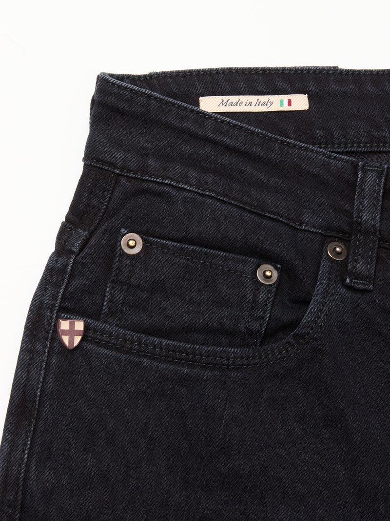Close-up of the upper part of Blue de Gênes Recco BlueBlk Jeans in blue-black denim, showing the front pocket, shield-shaped emblem, metal rivets, and a Made in Italy label with an Italian flag. These jeans feature a relaxed regular fit.