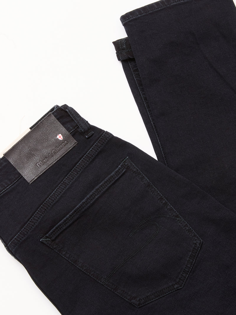 A pair of Recco BlueBlk Jeans by Blue de Gênes, crafted in dark blue-black Italian denim with a visible back pocket, black leather brand patch on the waistband, and tapered leg, is folded on a white surface.