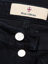 Close-up of Recco BlueBlk Jeans from Blue de Gênes in dark blue-black denim, featuring metal buttons with a shield logo and a beige Blue de Gênes label topped with a red cross emblem.