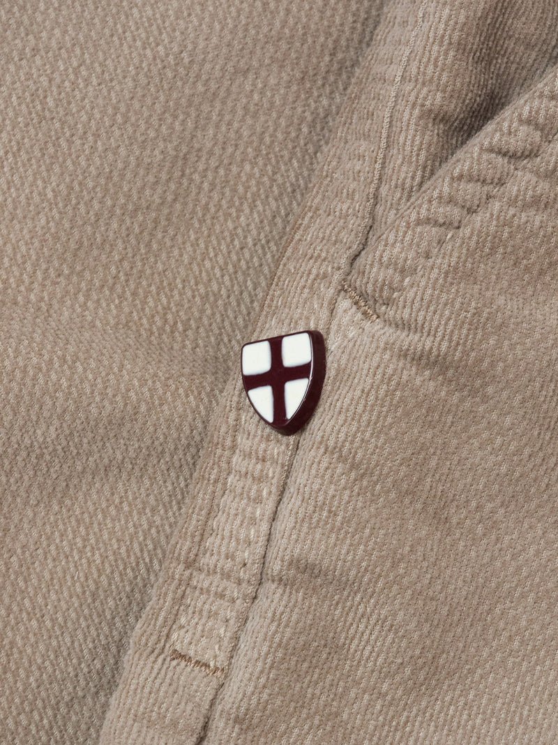 A small white shield pin with a red cross accents the pocket seam of the tan BGpaulo Ottawa Trousers - Fungi by Blue de Gênes, adding a subtle touch to these classic Italian-made chinos.