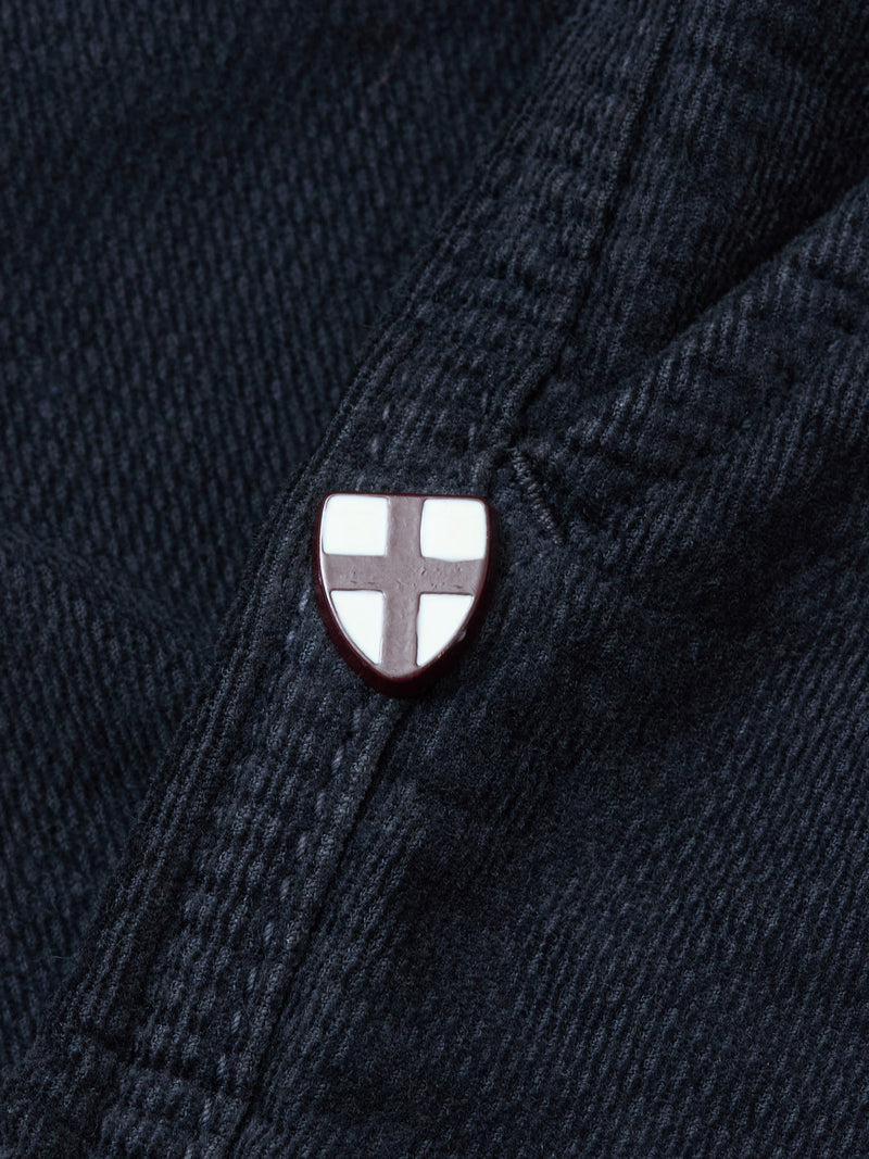 A close-up of dark denim with a shield-shaped enamel pin, white with a red cross—perfectly paired with Blue de Gênes BGpaulo Ottawa Trousers in Dark Navy for a refined, distinctive touch.