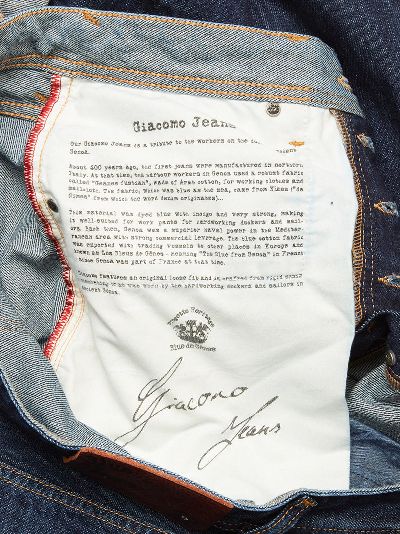 A close-up shows the inside pocket of Blue de Gênes Giacomo Dark Horse Jeans in denim blue, featuring a white label with brand history and signature. Copper rivets and red stitching highlight the quality rigid denim craftsmanship.