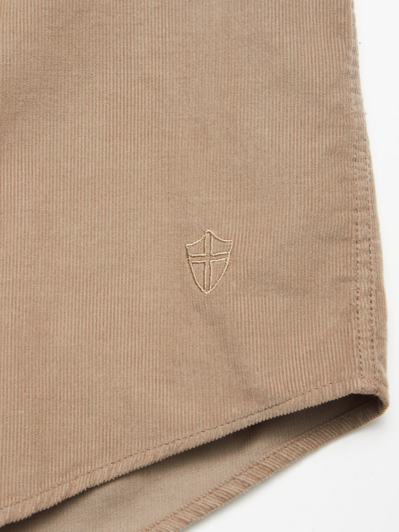 Close-up of the Blue de Gênes Zamboni Velu Shirt - Fungi, a garment-dyed baby corduroy piece with a small embroidered shield and cross near the hem, shown against a plain white background.
