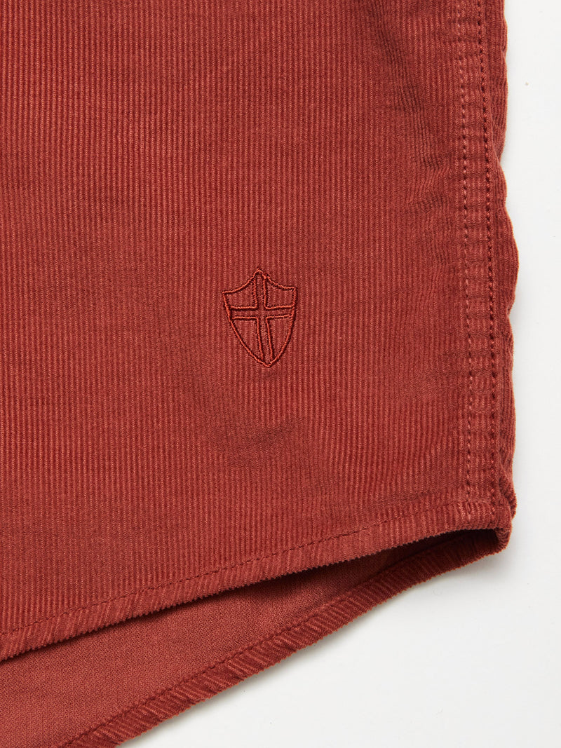 Close-up of the Zamboni Velu Shirt - Henna by Blue de Gênes, featuring red garment-dyed baby corduroy and an embroidered shield emblem near the hem, shown with fabric edge detail on a white background.