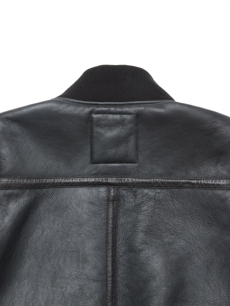 Close-up of the upper back of the Rufo Shearling Jacket - Scarab by Blue de Gênes, featuring black natural sheepskin, visible stitching, textured leather, a ribbed black collar, and a rectangular stitched patch.