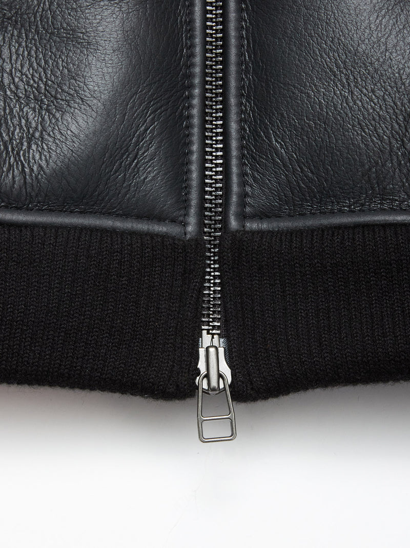 Close-up of the Rufo Shearling Jacket - Scarab by Blue de Gênes, featuring black natural sheepskin, a silver zipper, textured leather, and ribbed black fabric at the hem.