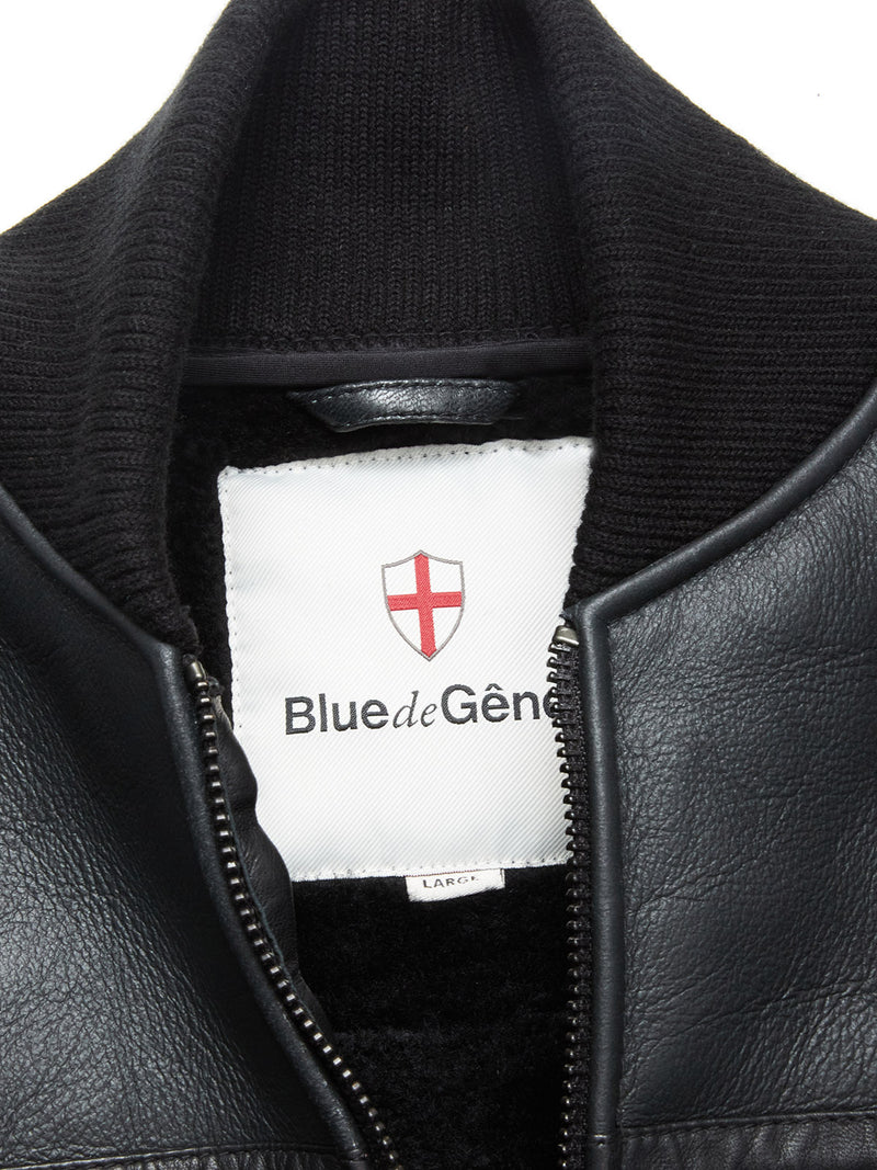 Close-up of the Rufo Shearling Jacket - Scarab by Blue de Gênes, featuring a black leather finish, ribbed collar, and partially unzipped to show a white Blue de Gênes label with a red shield emblem inside.