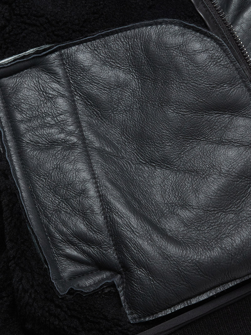 A close-up of the Blue de Gênes Rufo Shearling Jacket - Scarab shows its textured black leather, detailed stitching, and a partially unzipped front revealing a soft black fleece lining.