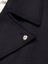 Close-up of the Blue de Gênes Shiba Melton Coat in Dark Navy, crafted from Italian Melton wool with a pointed lapel and metallic shield pin featuring a cross. A white label with black text appears in the background.