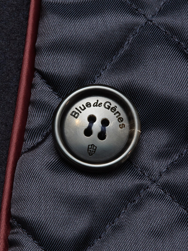 Close-up of a glossy button featuring the Blue de Gênes shield emblem, sewn onto dark navy Italian Melton wool with visible stitching and burgundy trim, highlighting the classic style of the Shiba Melton Coat by Blue de Gênes.