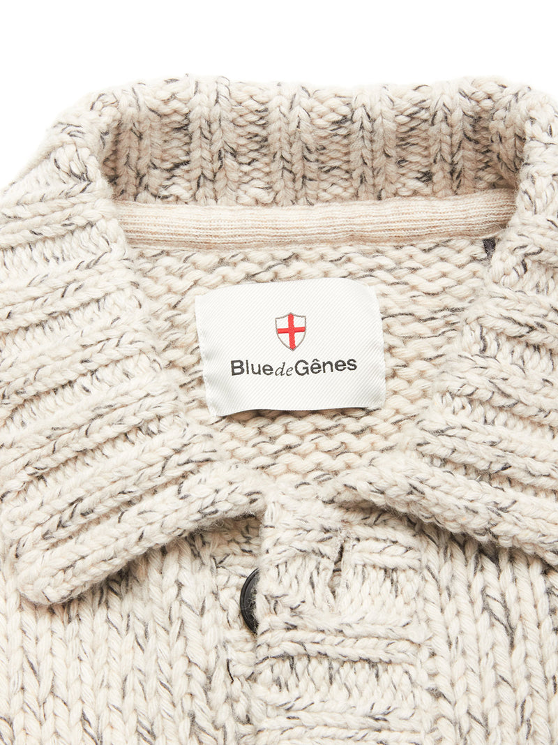 Close-up of the Sakura Knit Cardigan - Stone by Blue de Gênes, a chunky beige wool blend cardigan with a collar and a white label featuring the brand name and red cross emblem—ideal for layering on cooler days.