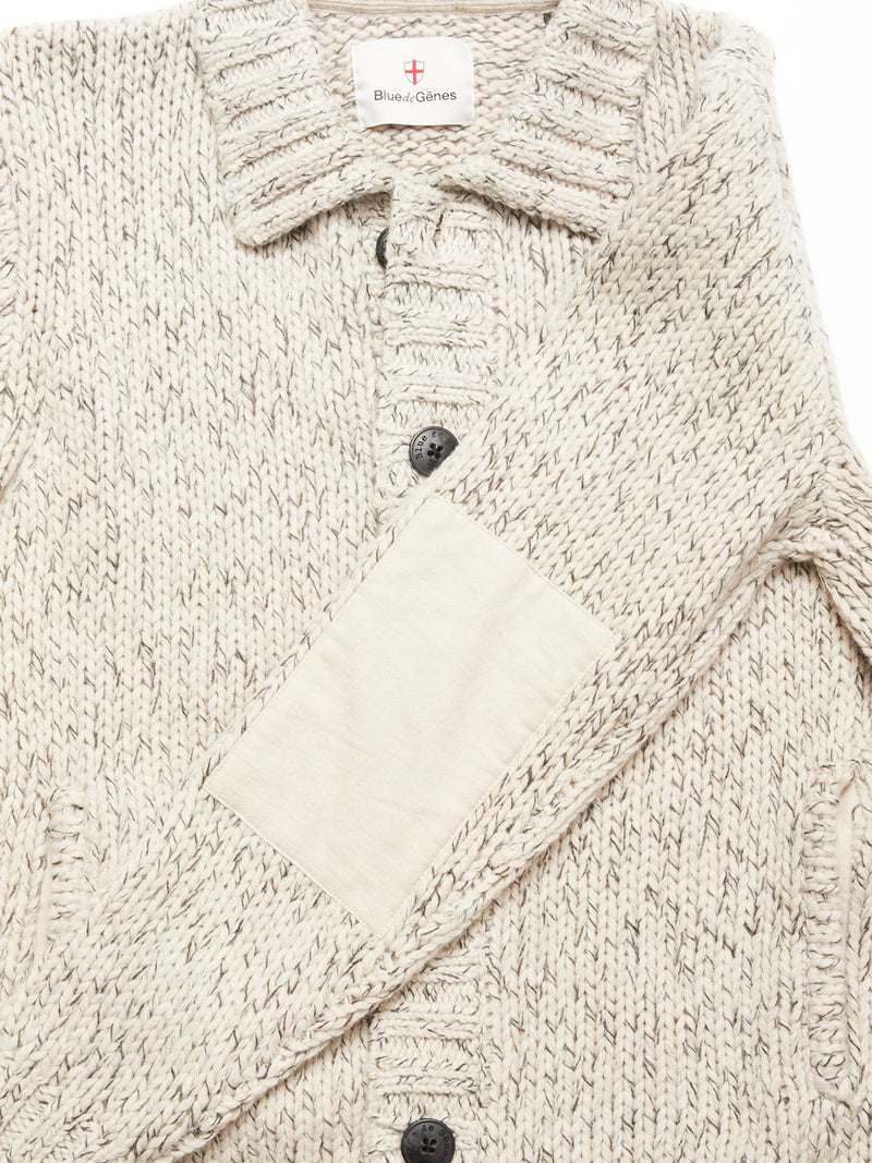 A close-up of the Sakura Knit Cardigan - Stone by Blue de Gênes, a beige speckled wool blend with dark buttons, chunky knit texture, collar, sleeve patch, and the Blue de Gênes label visible at the top.