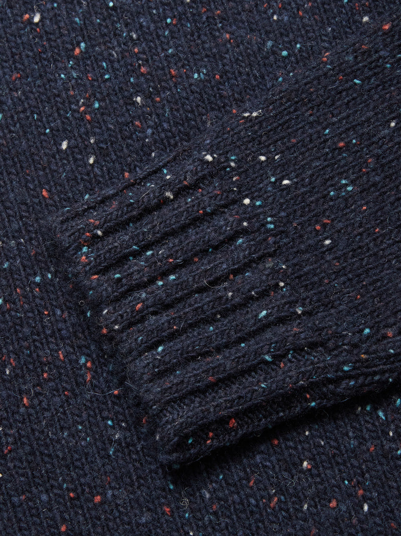 A close-up of the Blue de Gênes Honshu Crewneck Knit in navy, featuring colorful red, white, and blue speckles. The image highlights a ribbed cuff atop the cozy lambswool blend sweater.