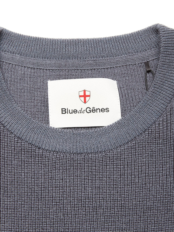 Close-up of the neckline on the Porto Knit - Dark Slate sweater by Blue de Gênes, featuring textured blue knit, a crewneck design, and a white label with Blue de Gênes branding and a red-and-white shield logo.