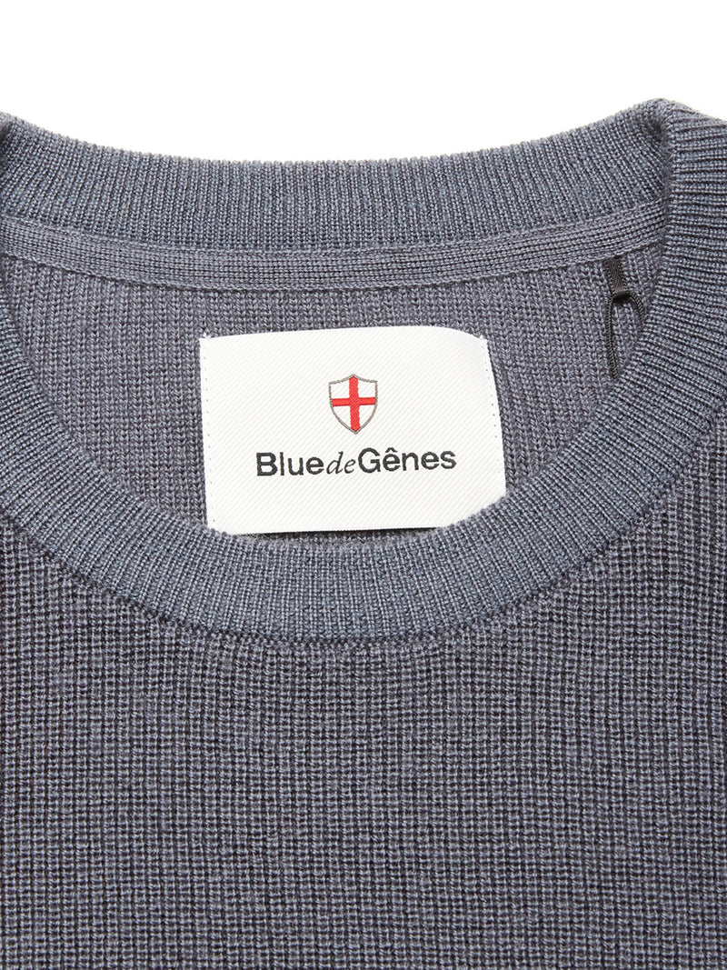 Close-up of the neckline on the Porto Knit - Dark Slate sweater by Blue de Gênes, featuring textured blue knit, a crewneck design, and a white label with Blue de Gênes branding and a red-and-white shield logo.