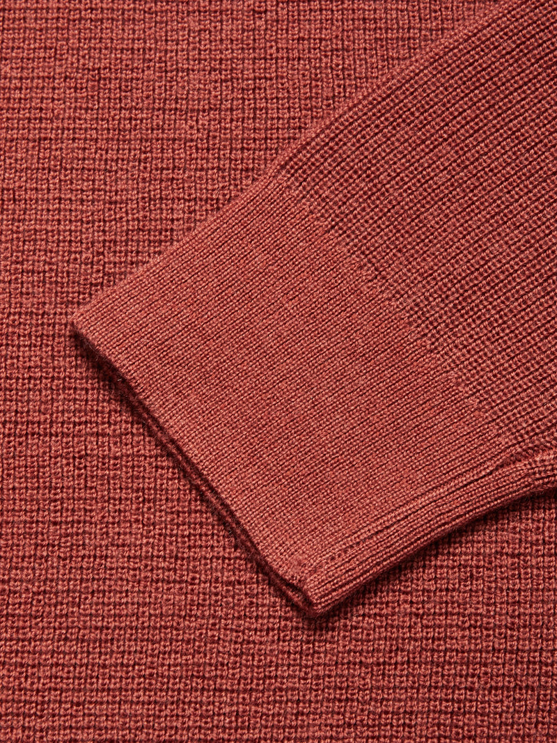 A close-up of the Blue de Gênes Porto Knit - Henna crewneck sweater shows the sleeve cuff and body, highlighting its rich, textured reddish-brown knit pattern.