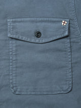 Close-up of a dark slate cotton Nagano Overshirt pocket by Blue de Gênes, featuring a flap with a black button and a small white patch with a red X sewn near the top right corner of the garment-dyed pocket flap.