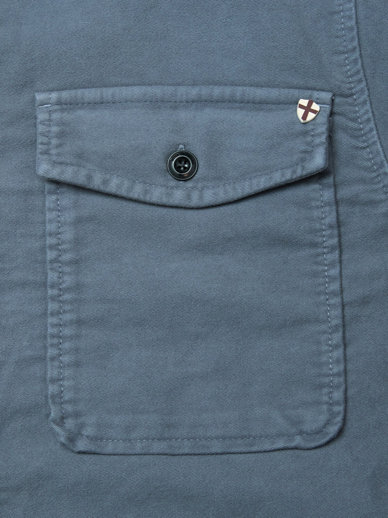 Close-up of a dark slate cotton Nagano Overshirt pocket by Blue de Gênes, featuring a flap with a black button and a small white patch with a red X sewn near the top right corner of the garment-dyed pocket flap.