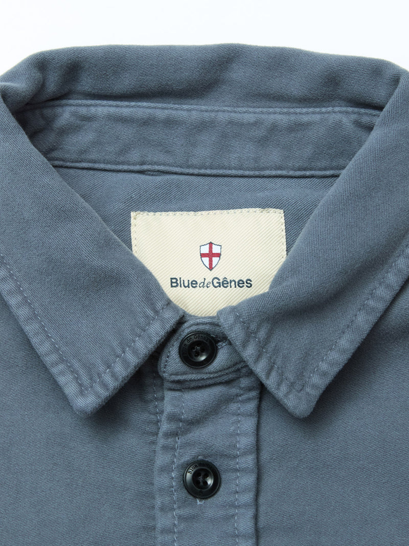 A close-up of the Nagano Overshirt - Dark Slate by Blue de Gênes, featuring black buttons and an inner collar label with the brand’s name and a red and white shield logo above the text.
