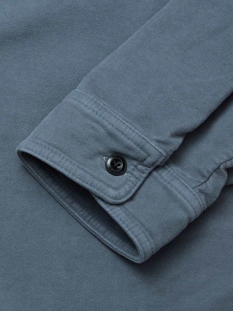 A close-up of a Blue de Gênes Nagano Overshirt - Dark Slate cuff with a black button, showcasing the garment dyed cotton Moleskin fabric texture and detailed stitching.