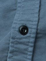 Close-up of a dark blue button sewn onto blue fabric from the Blue de Gênes Nagano Overshirt - Dark Slate. Visible stitching lines the placket, and small text is inscribed around the button’s edge.