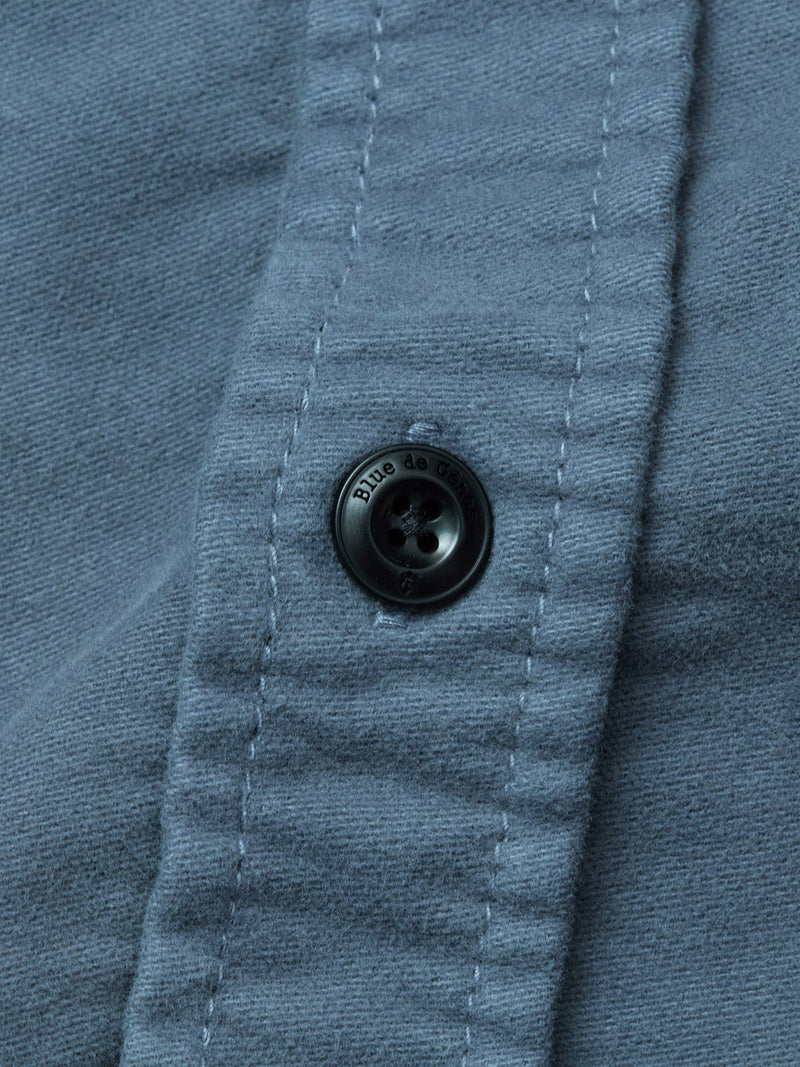 Close-up of a dark blue button sewn onto blue fabric from the Blue de Gênes Nagano Overshirt - Dark Slate. Visible stitching lines the placket, and small text is inscribed around the button’s edge.