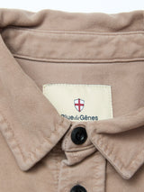 A close-up of the Nagano Overshirt - Fungi by Blue de Gênes, crafted in beige cotton moleskin with black buttons. The collar label features a small shield with a red cross and the Blue de Gênes brand name.