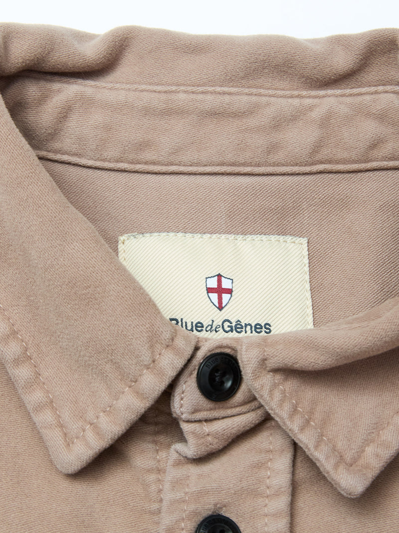 A close-up of the Nagano Overshirt - Fungi by Blue de Gênes, crafted in beige cotton moleskin with black buttons. The collar label features a small shield with a red cross and the Blue de Gênes brand name.
