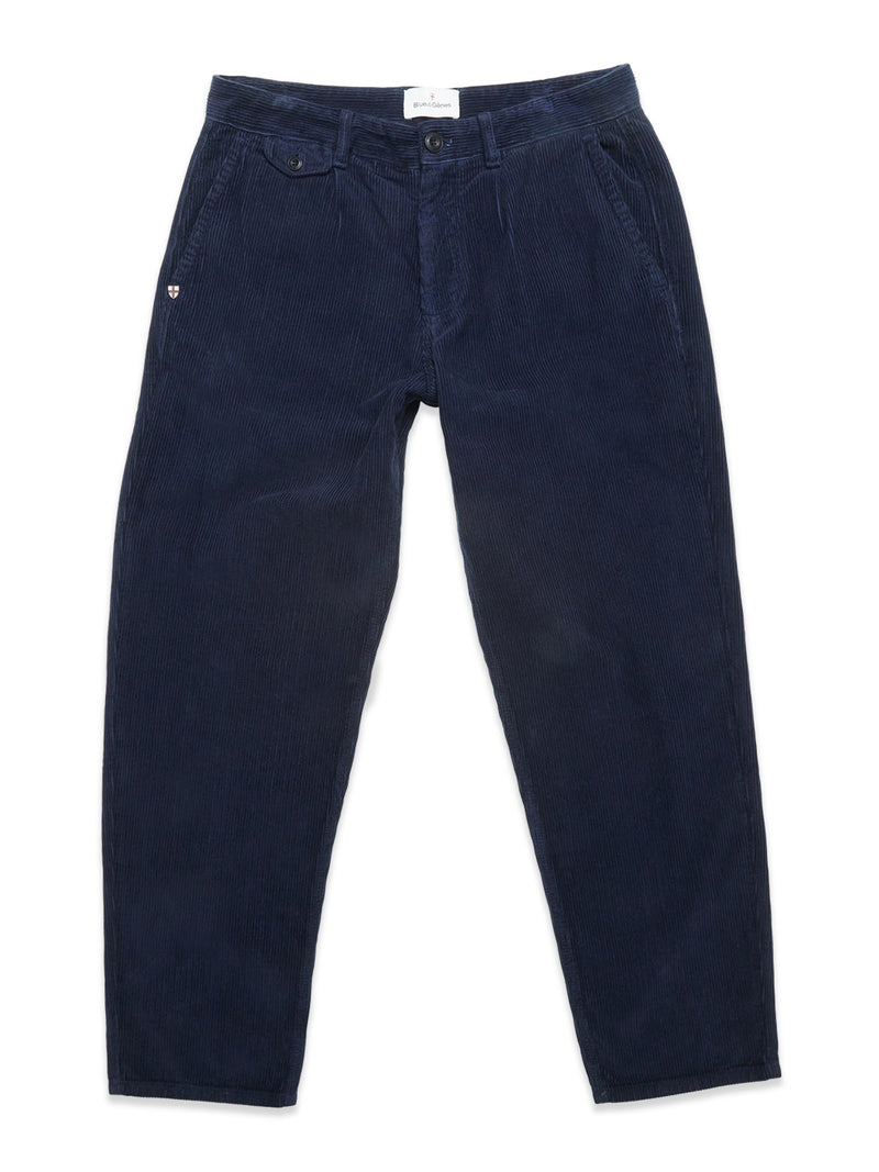 The Blue de Gênes Etsu Cord Chino in navy is a pair of garment-dyed, relaxed fit corduroy trousers with button and zip closure, slanted front pockets, and a logo patch on the left pocket, shown laid flat on white.