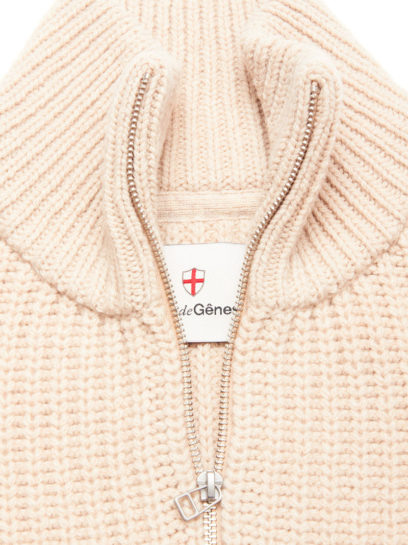 Close-up of the Yomaha Halfzip Knit - Fungi by Blue de Gênes, a cream chunky knit sweater with a ribbed half-zip collar, partially unzipped to show a white label with a red shield logo and text.