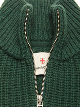 Close-up of the Yomaha Halfzip Knit - Scarab by Blue de Gênes: a green chunky wool blend jumper with a silver half-zip partially open, revealing a white label with red cross emblem and partial text beneath the collar.