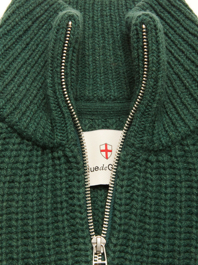 Close-up of the Yomaha Halfzip Knit - Scarab by Blue de Gênes: a green chunky wool blend jumper with a silver half-zip partially open, revealing a white label with red cross emblem and partial text beneath the collar.