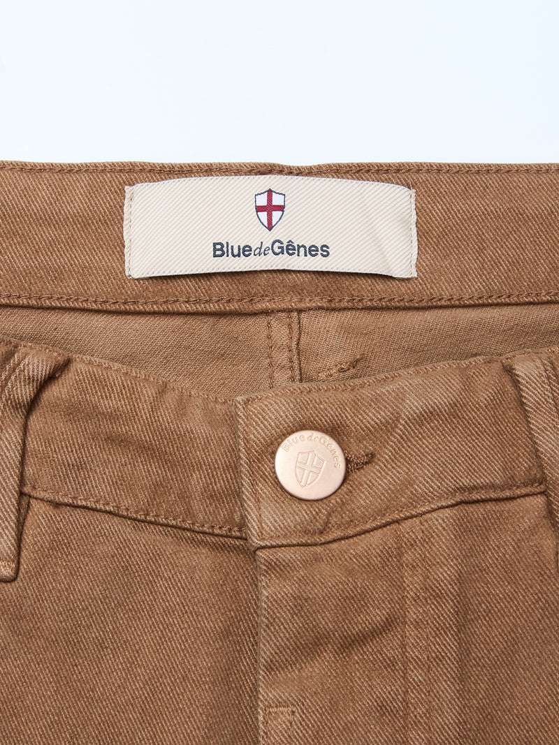 Close-up of the Vinci Iro Pant - Chocolate by Blue de Gênes, showing the comfort stretch waistband with a metal button embossed with a shield and a white label featuring a red cross and Blue de Gênes text.
