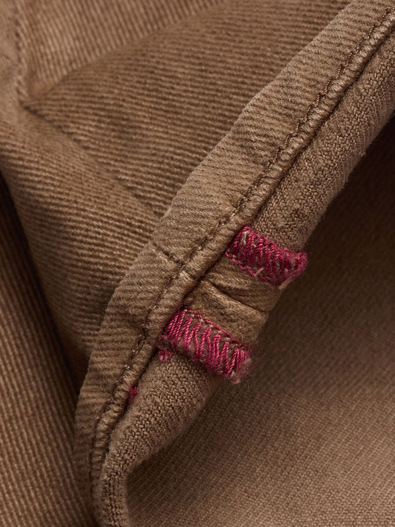 Close-up of the Vinci Iro Pant - Chocolate by Blue de Gênes, showing brown fabric with visible stitching and two buttonholes reinforced in pink thread. The texture and weave are inspired by regular fit jeans.