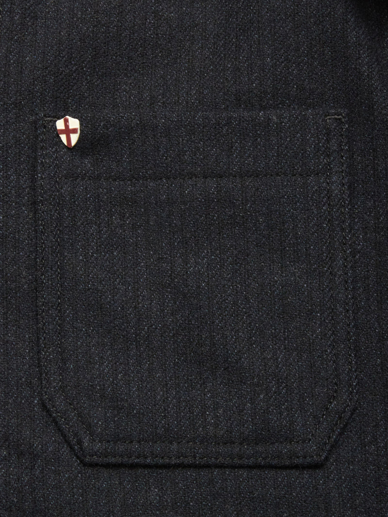 Close-up of a dark denim pocket with a small pin, alongside the Blue de Gênes Vincenzo Solid Jacket - Dark Navy, featuring a distinctive white shield design with red cross.