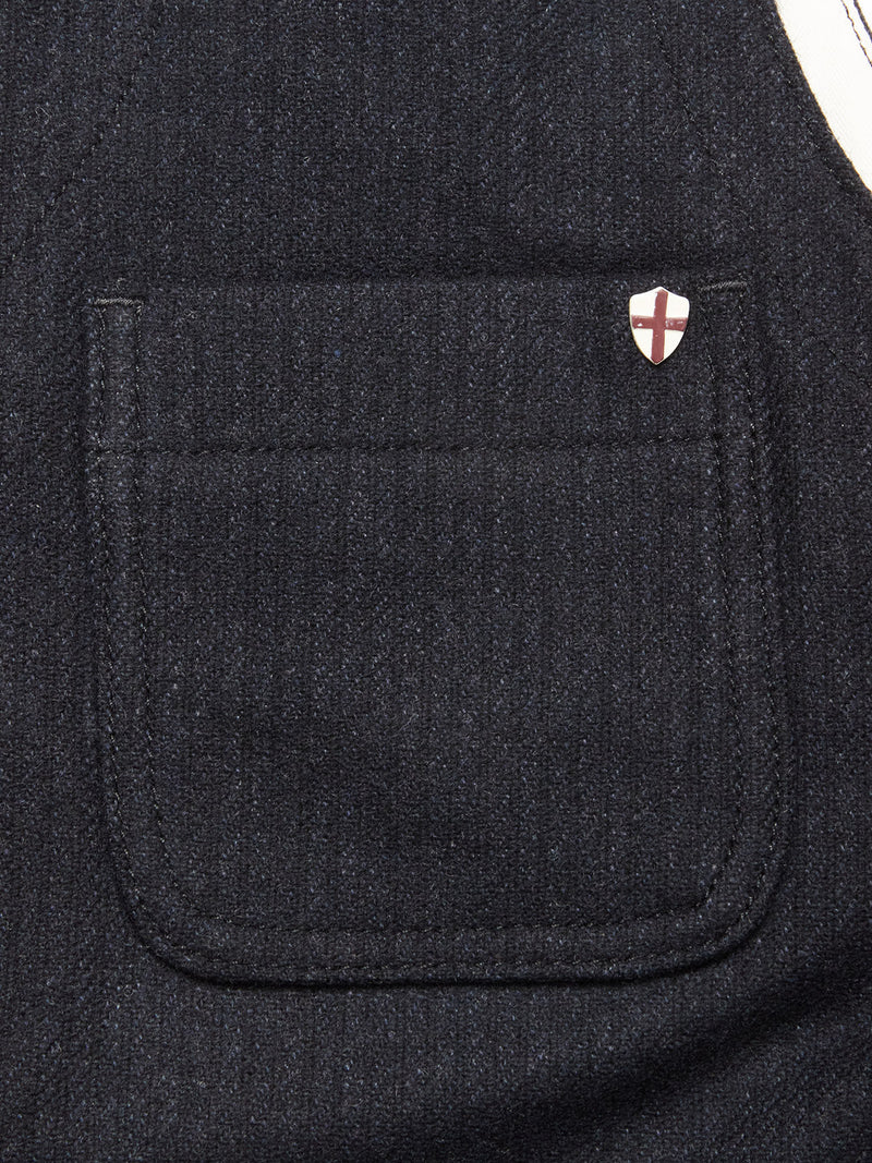 Close-up of a dark fabric pocket on the Maurice Solid Gilet - Dark Navy by Blue de Gênes, an Italian wool blend piece, with a red-cross shield pin on the top right corner of this tailored layer.