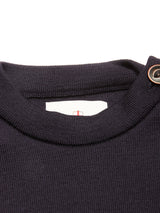 Close-up of the Toyama Knit L/S sweater by Blue de Gênes in dark navy, showing its ribbed collar with a button detail and a partially visible white tag featuring red and gray lines inside the neckline.