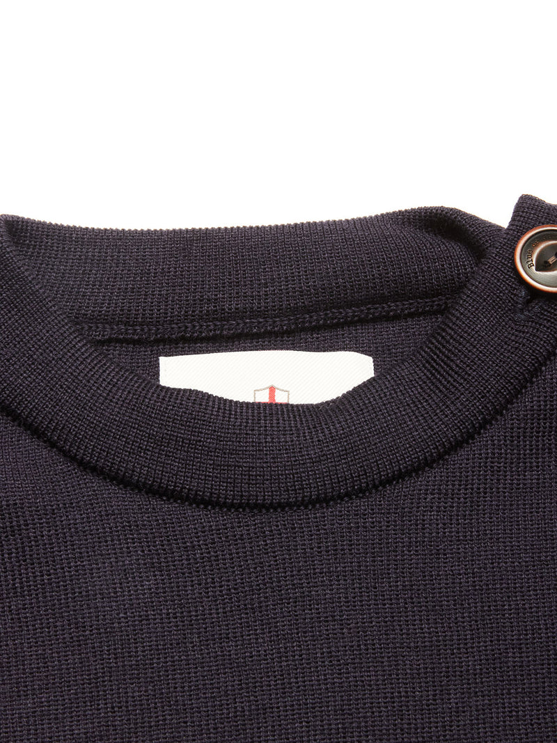 Close-up of the Toyama Knit L/S sweater by Blue de Gênes in dark navy, showing its ribbed collar with a button detail and a partially visible white tag featuring red and gray lines inside the neckline.